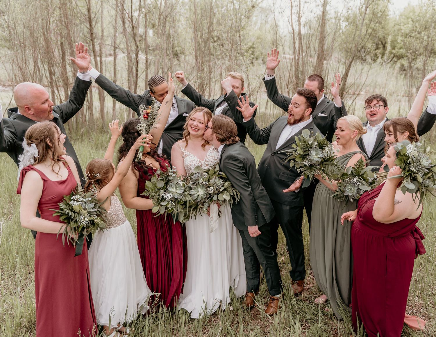 Wedding party celebrates joyfully outdoors with raised arms and greenery bouquets in burgundy and gray attire.