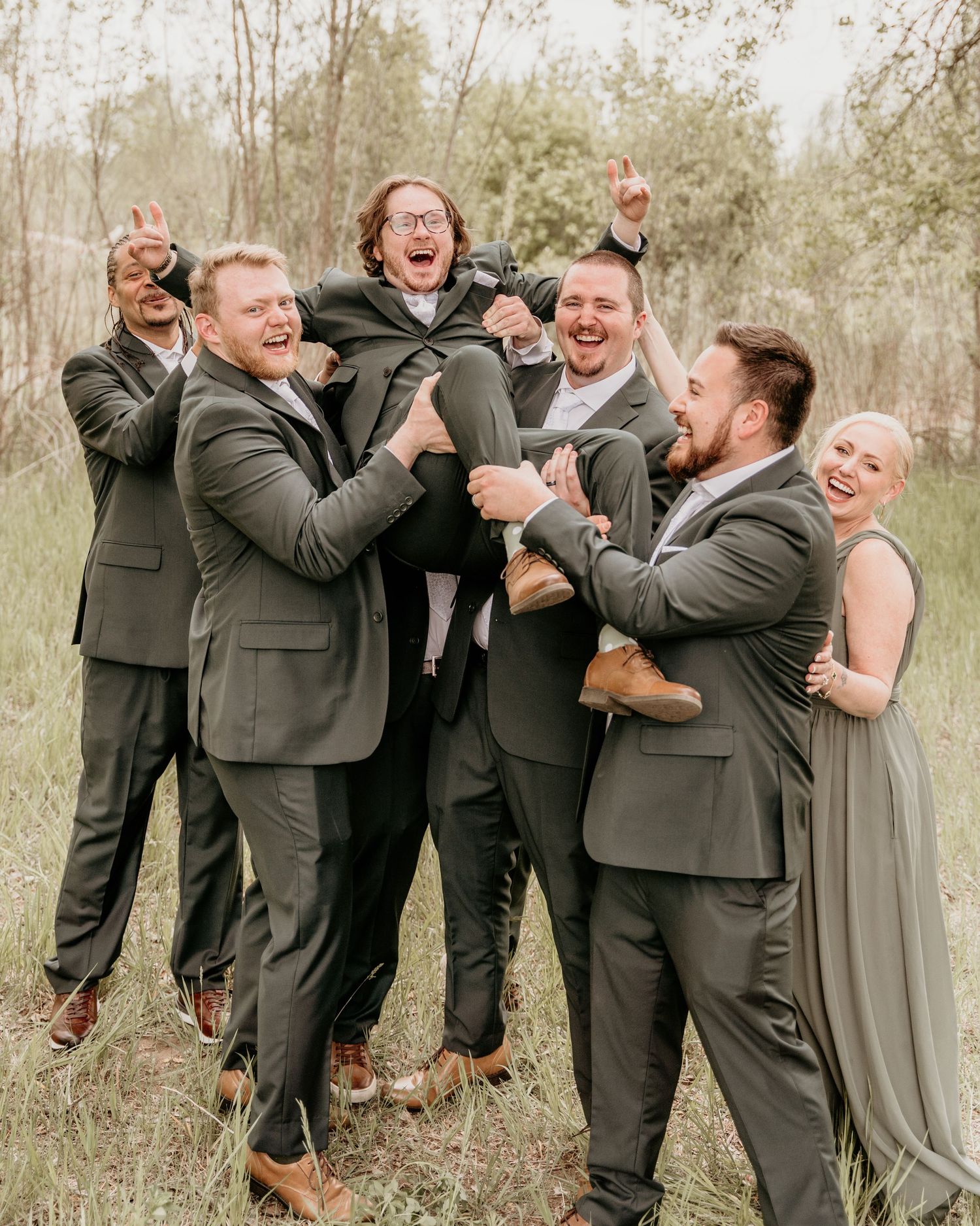 Wedding party in gray suits lifts and celebrates joyfully together in an outdoor nature setting.