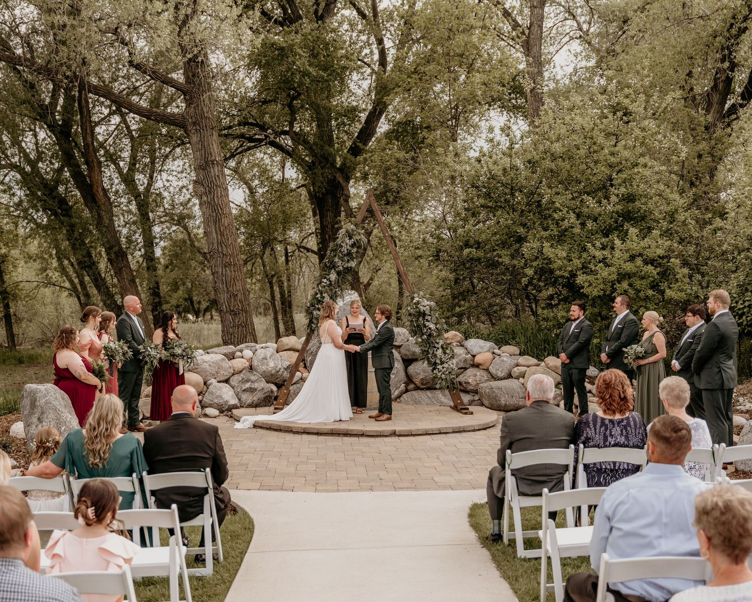 An outdoor wedding ceremony in a wooded setting with white chairs and a stone-lined pathway.