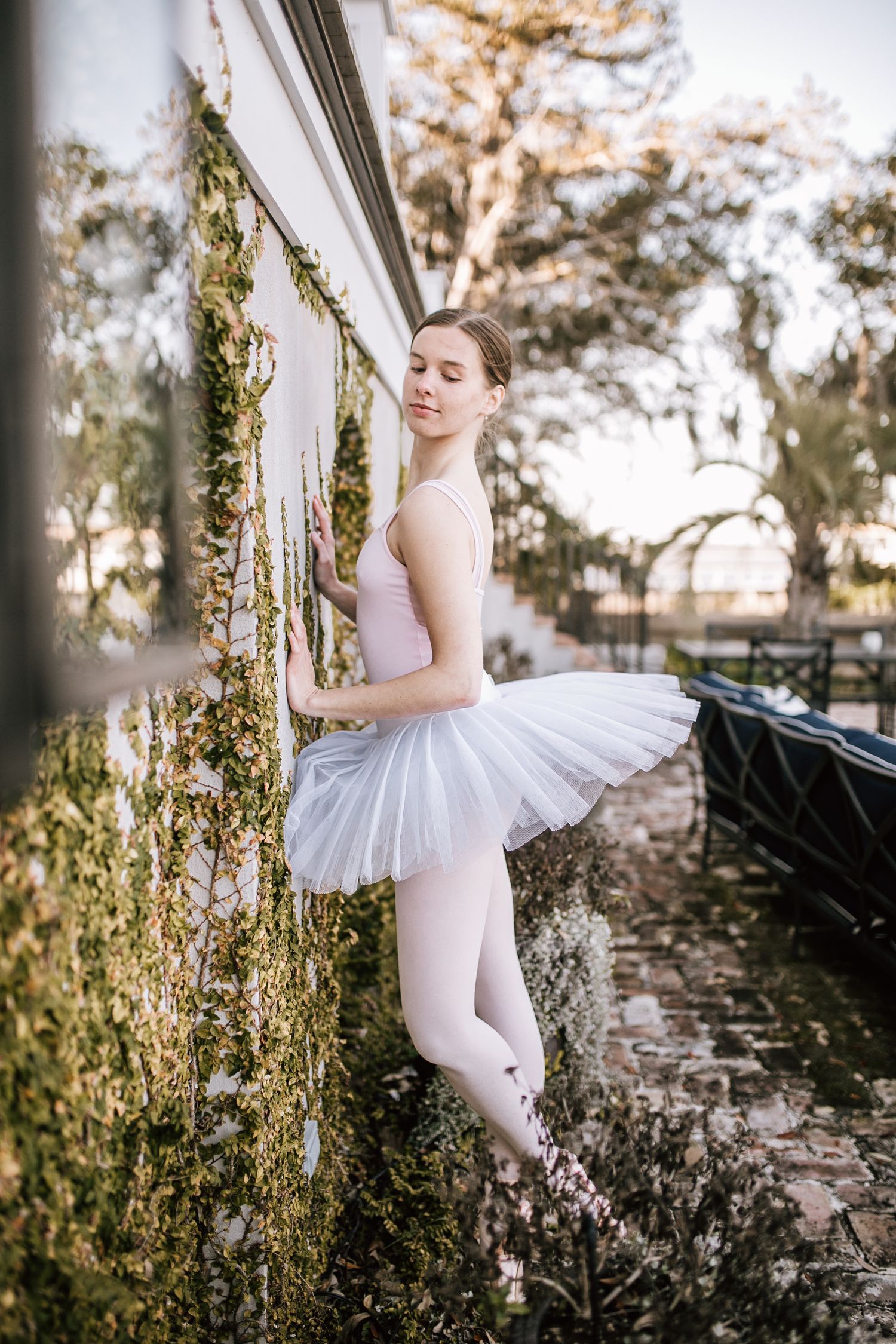 A graceful ballerina in white tutu and pink leotard poses en pointe against an ivy-covered wall outdoors.