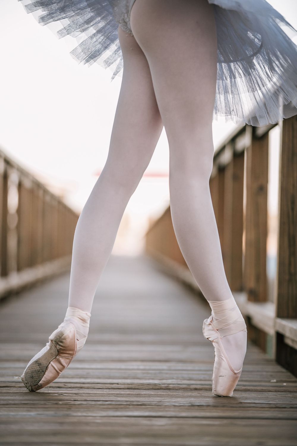 Close-up of beautifully arched ballet dancer's feet in pink pointe shoes and gray tutu on a wooden bridge in dance photography on SC dock.