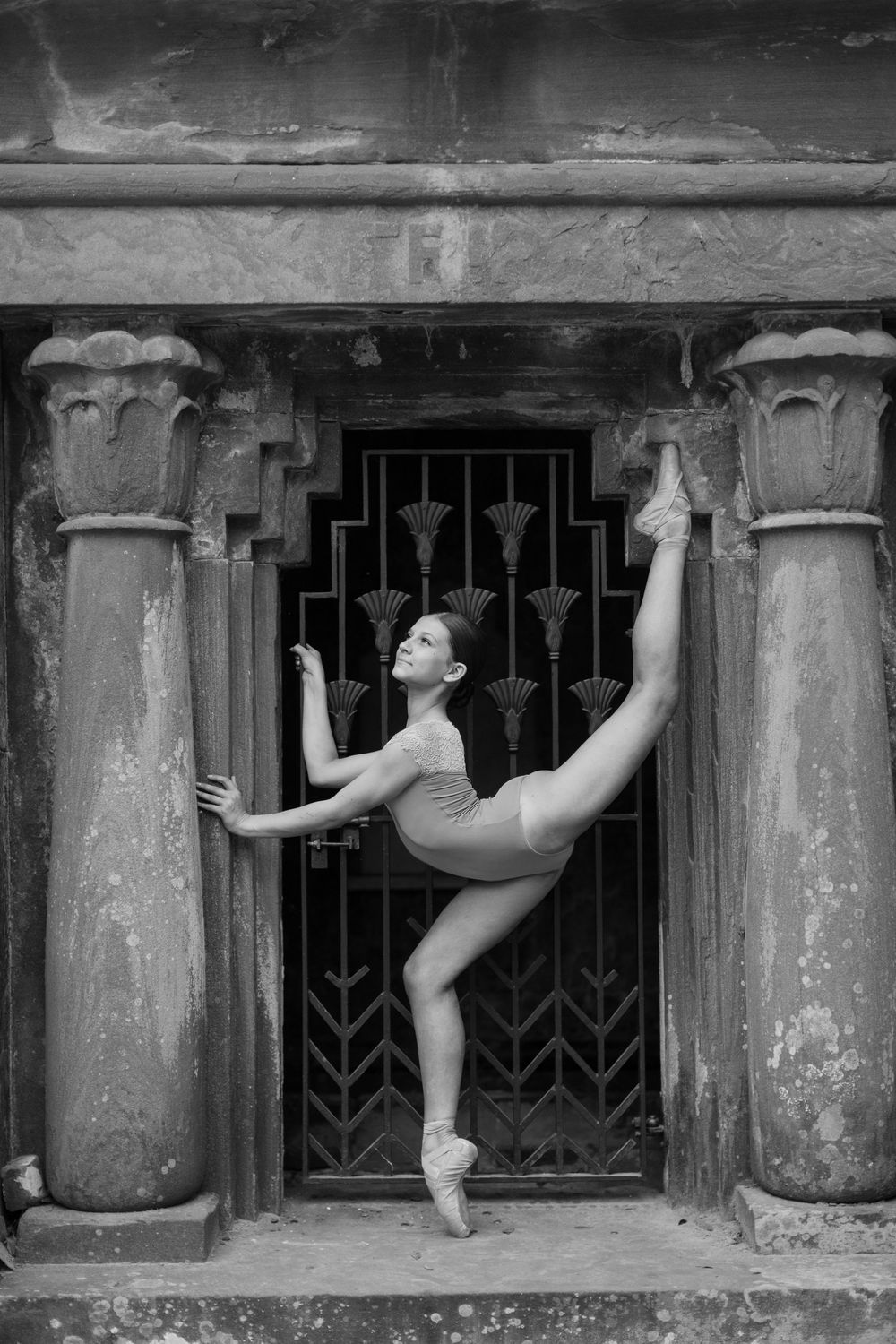 Black and white photo of a ballerina in pointe shoes performing a high leg extension pose before ornate stone columns.