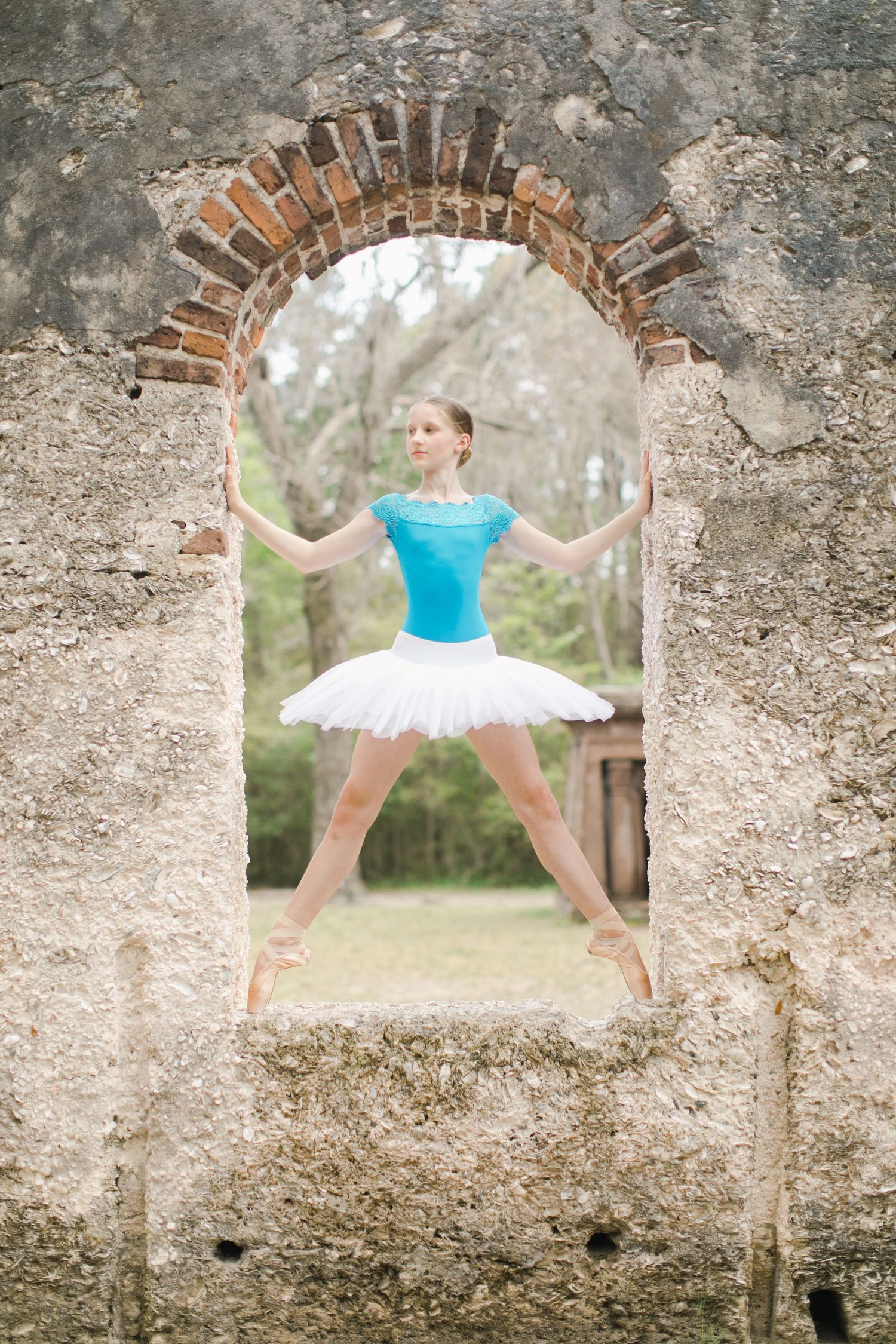 Ballerina in blue top and white tutu poses gracefully in a rustic stone archway surrounded by trees.