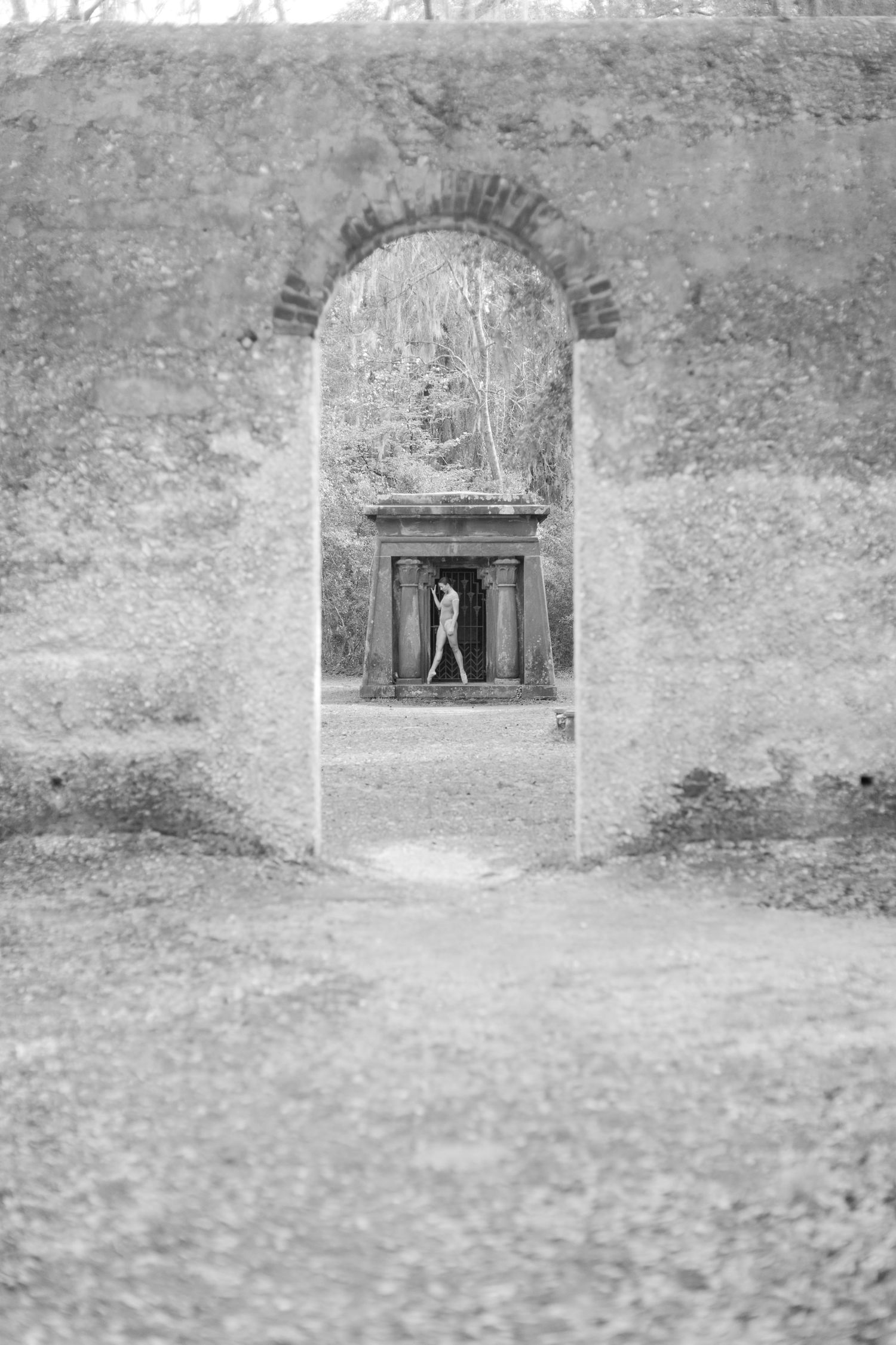 Black and white photo of a person standing in a stone archway leading to an ornate monument surrounded by bare trees at Chapel of Ease.