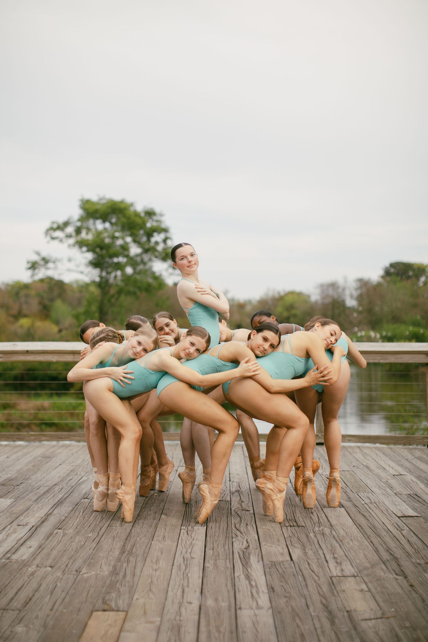 Group of ballet dancers in teal leotards posing together on wooden deck outdoors.
