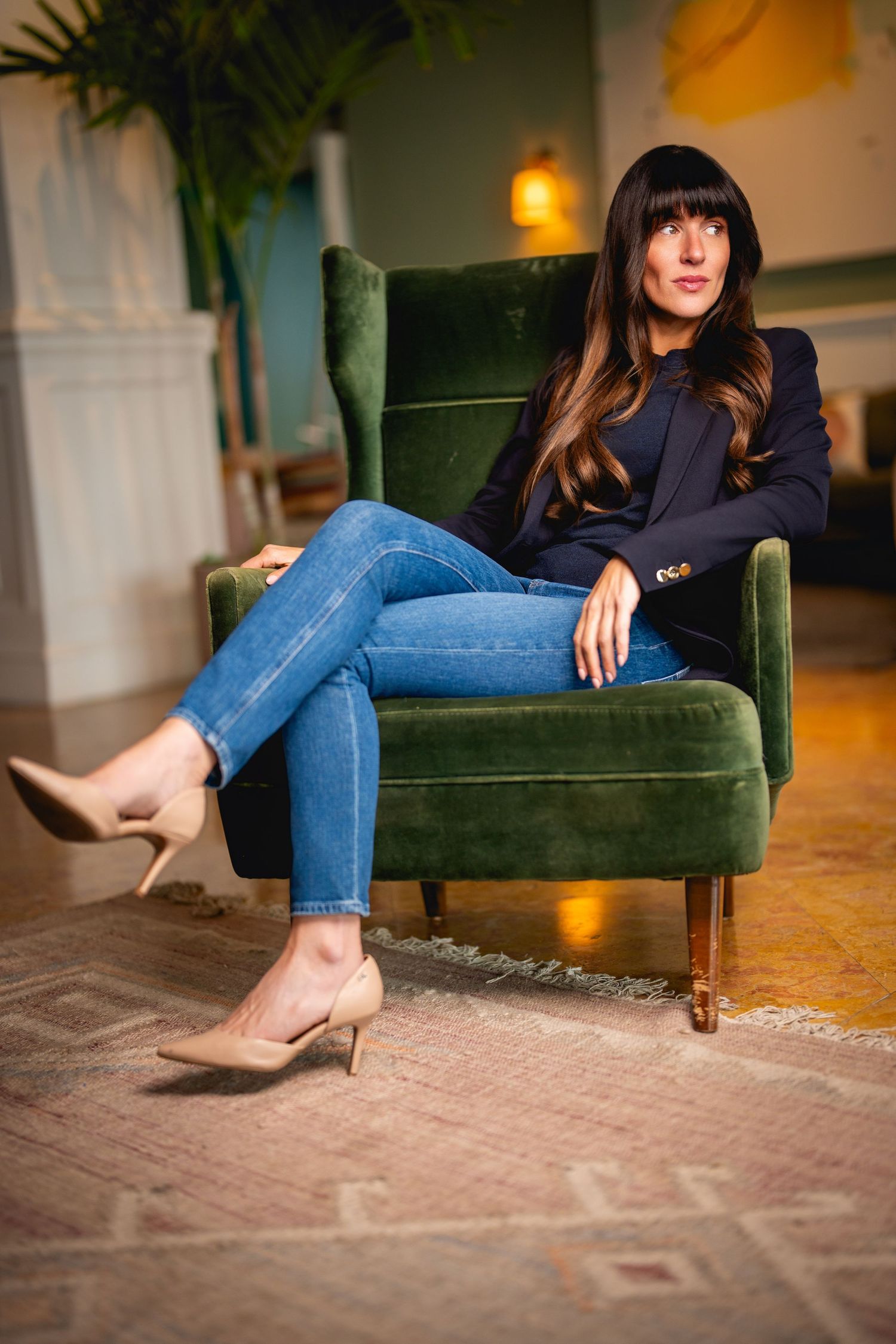 Stylish woman in blue jeans and navy top lounges in emerald green velvet armchair with beige heels.