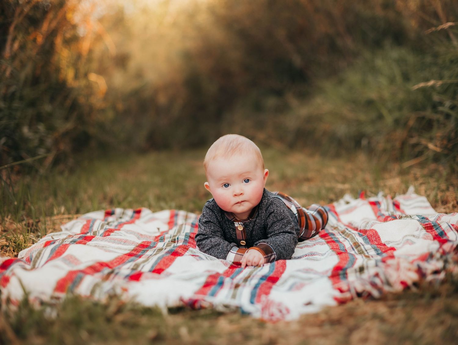 Baby in grey sweater on striped blanket outdoors during sunset.