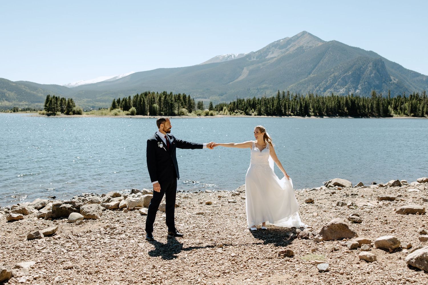 A couple in wedding attire holds hands while standing by a Colorado mountain lake with peaks and forest in the background.
