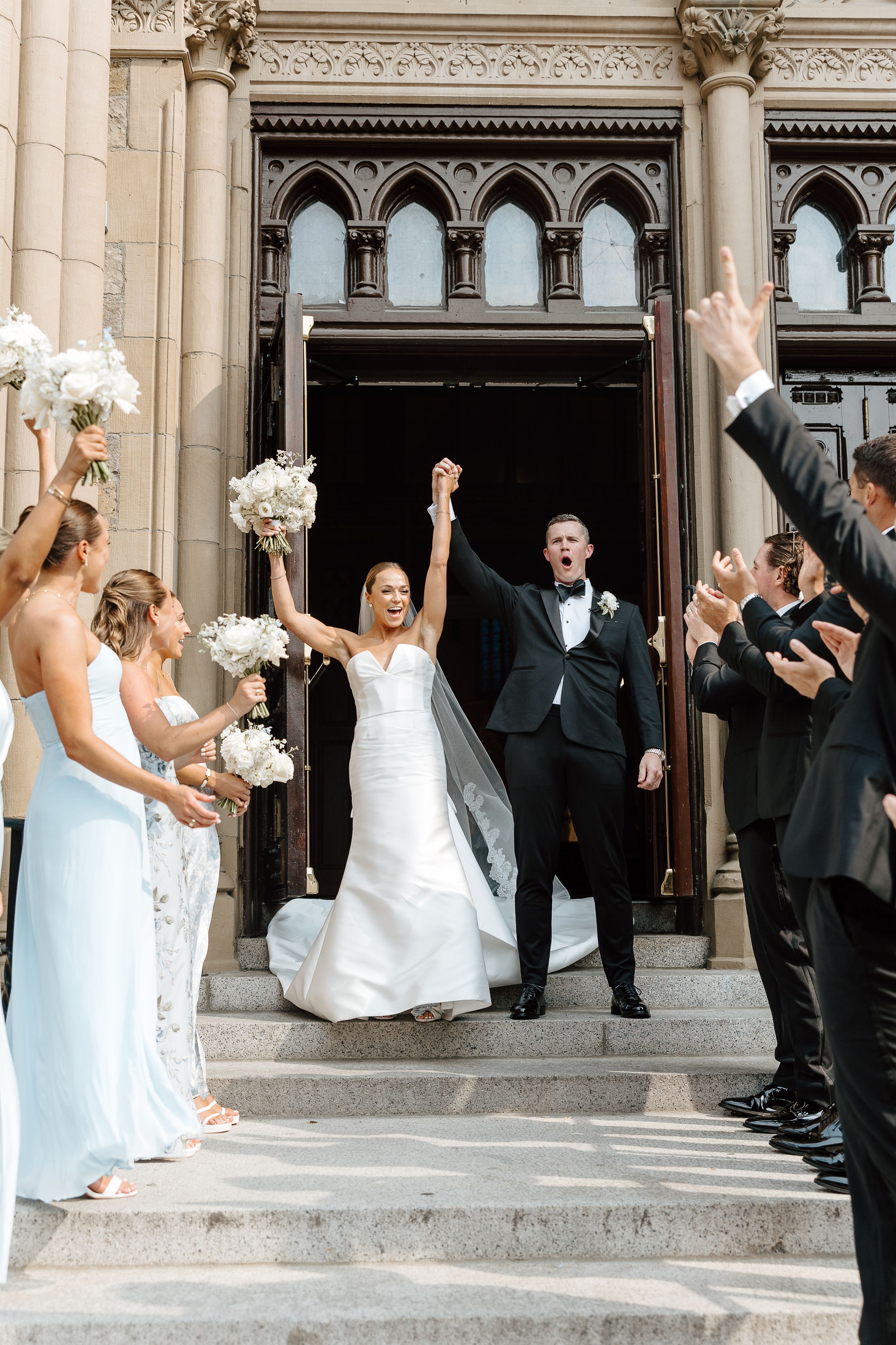 Wedding party celebrating with raised arms on church steps in formal attire.
