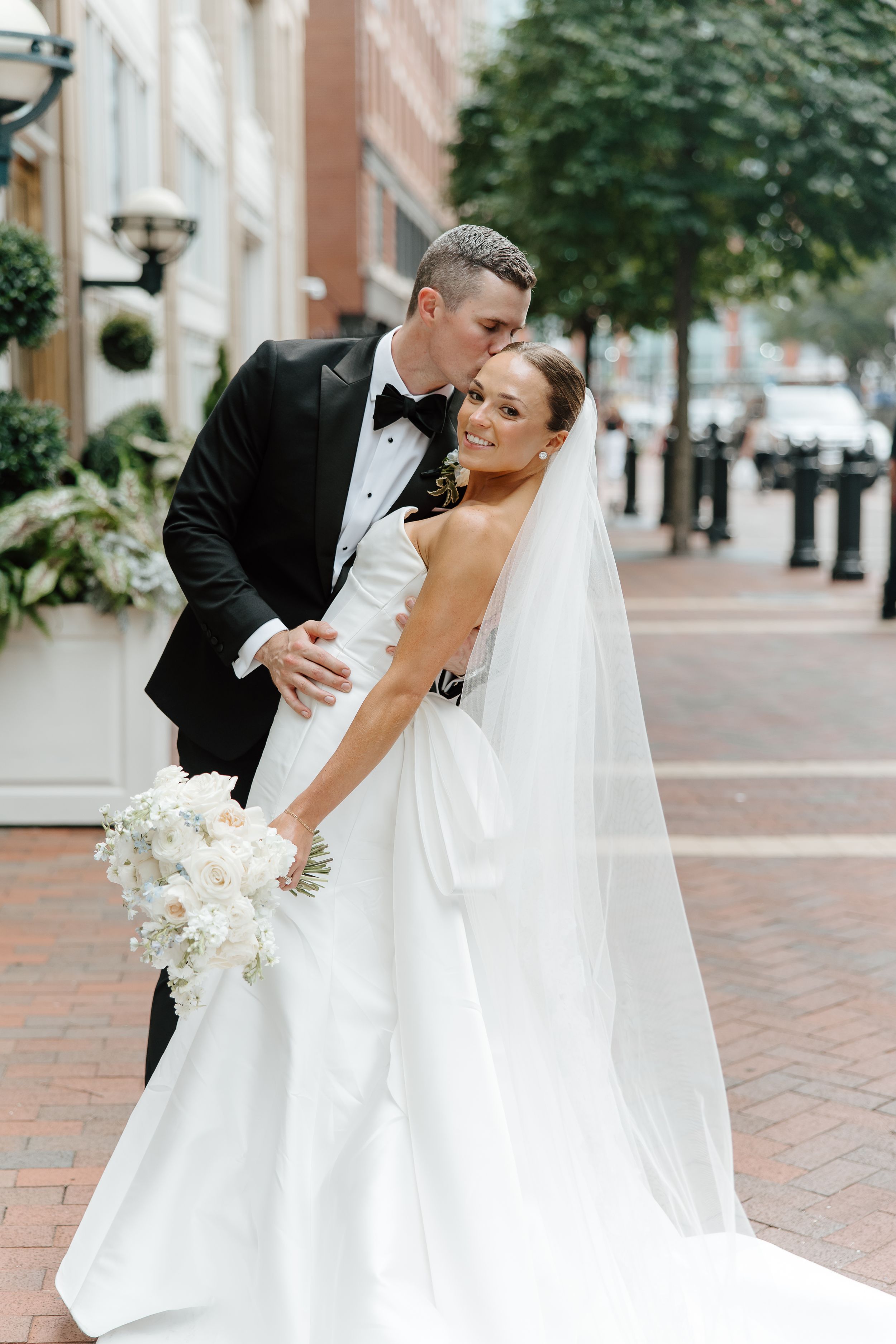 Elegant wedding couple poses on brick sidewalk with classic architecture backdrop.