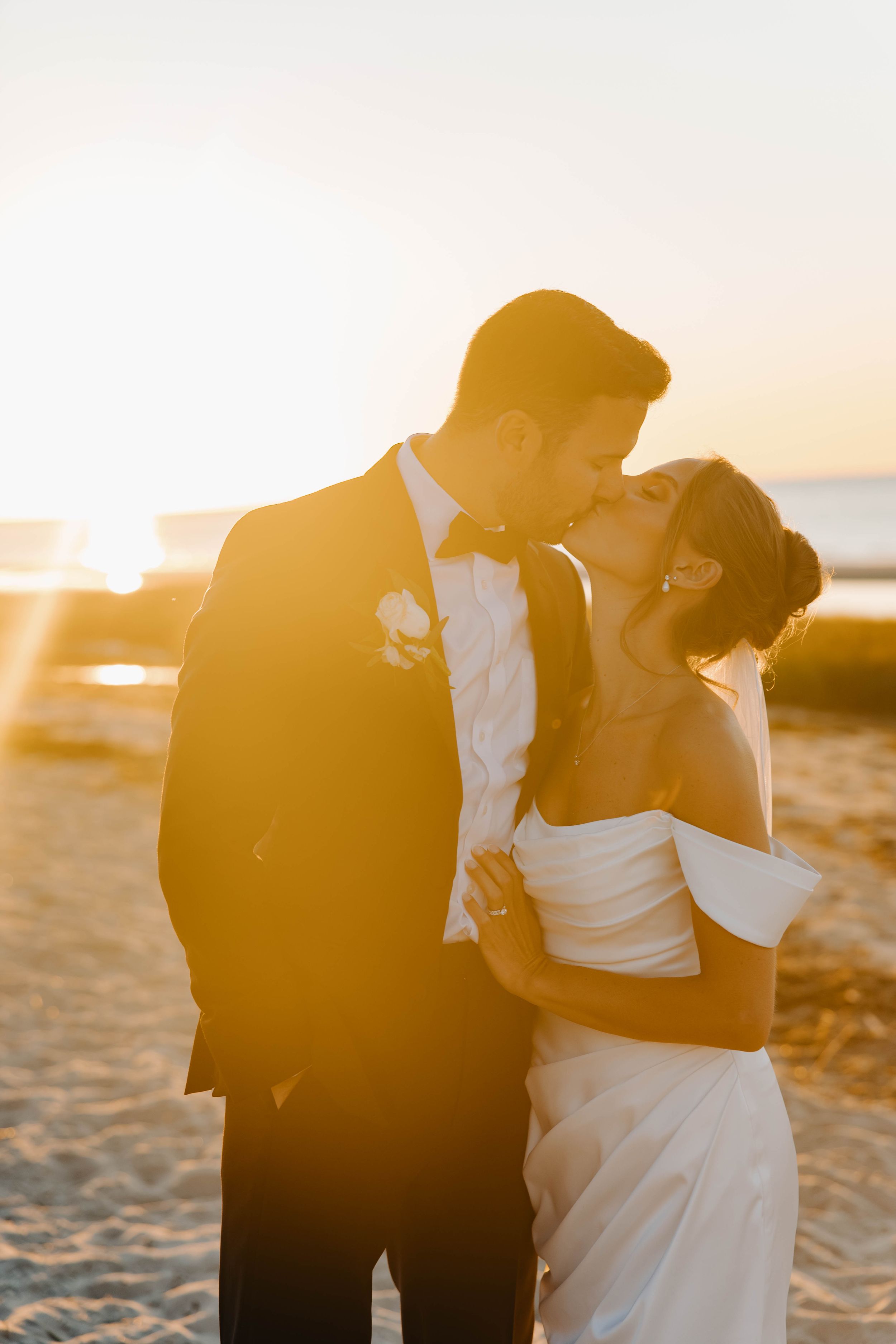 Bride and groom share a romantic sunset kiss on the beach, with golden light glowing behind them.