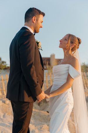A bride and groom hold hands on a sandy beach at golden hour, smiling at each other in wedding attire.