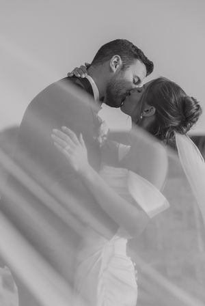 Black and white wedding photo of bride and groom sharing a romantic kiss, with veil flowing around them.