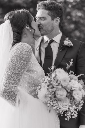 Black and white photo of a bride and groom sharing a kiss, bride holding a bouquet of roses on their wedding day.