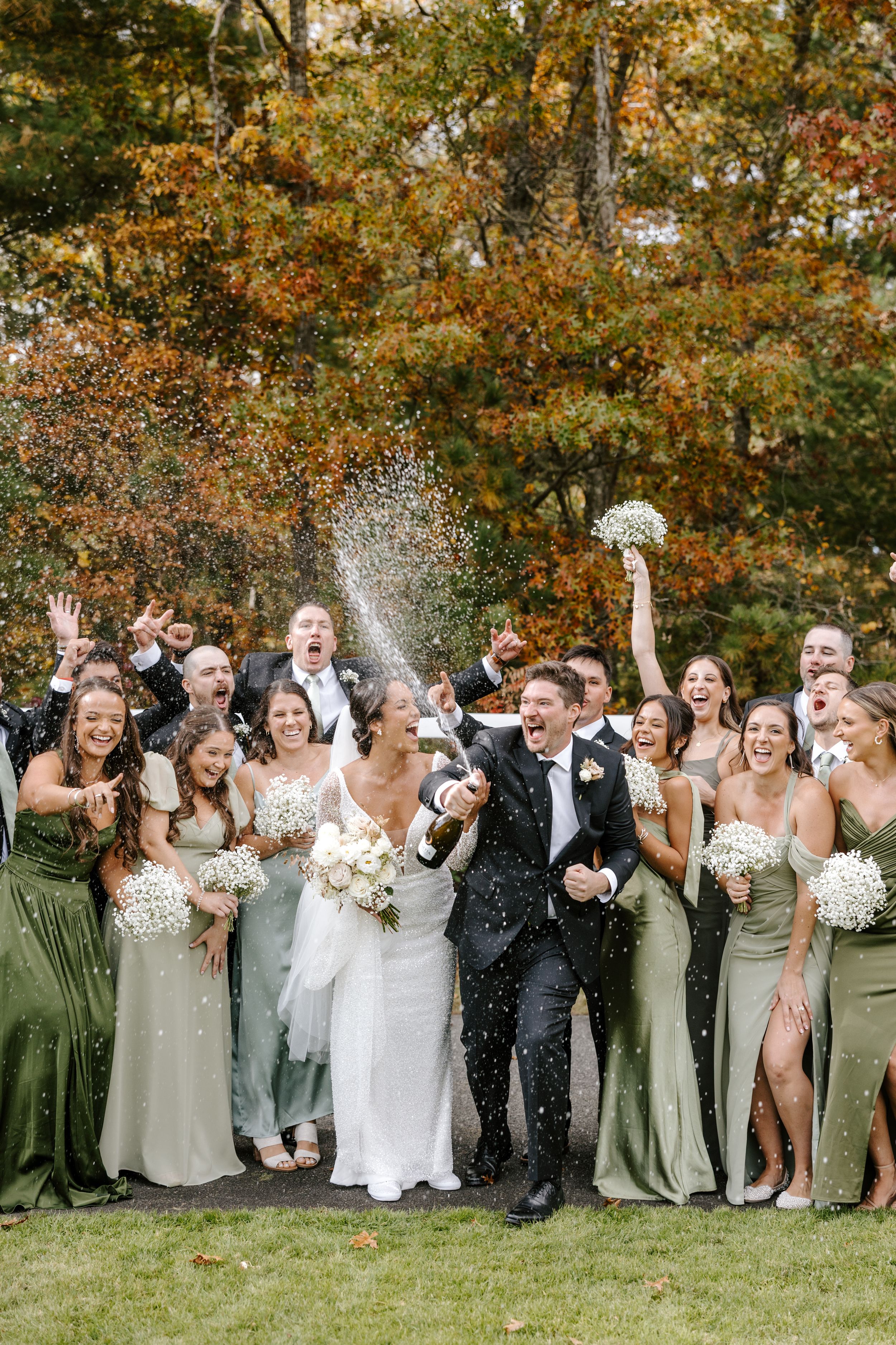 Wedding party celebrates with champagne spray outdoors, bridesmaids in sage green gowns, fall foliage backdrop.
