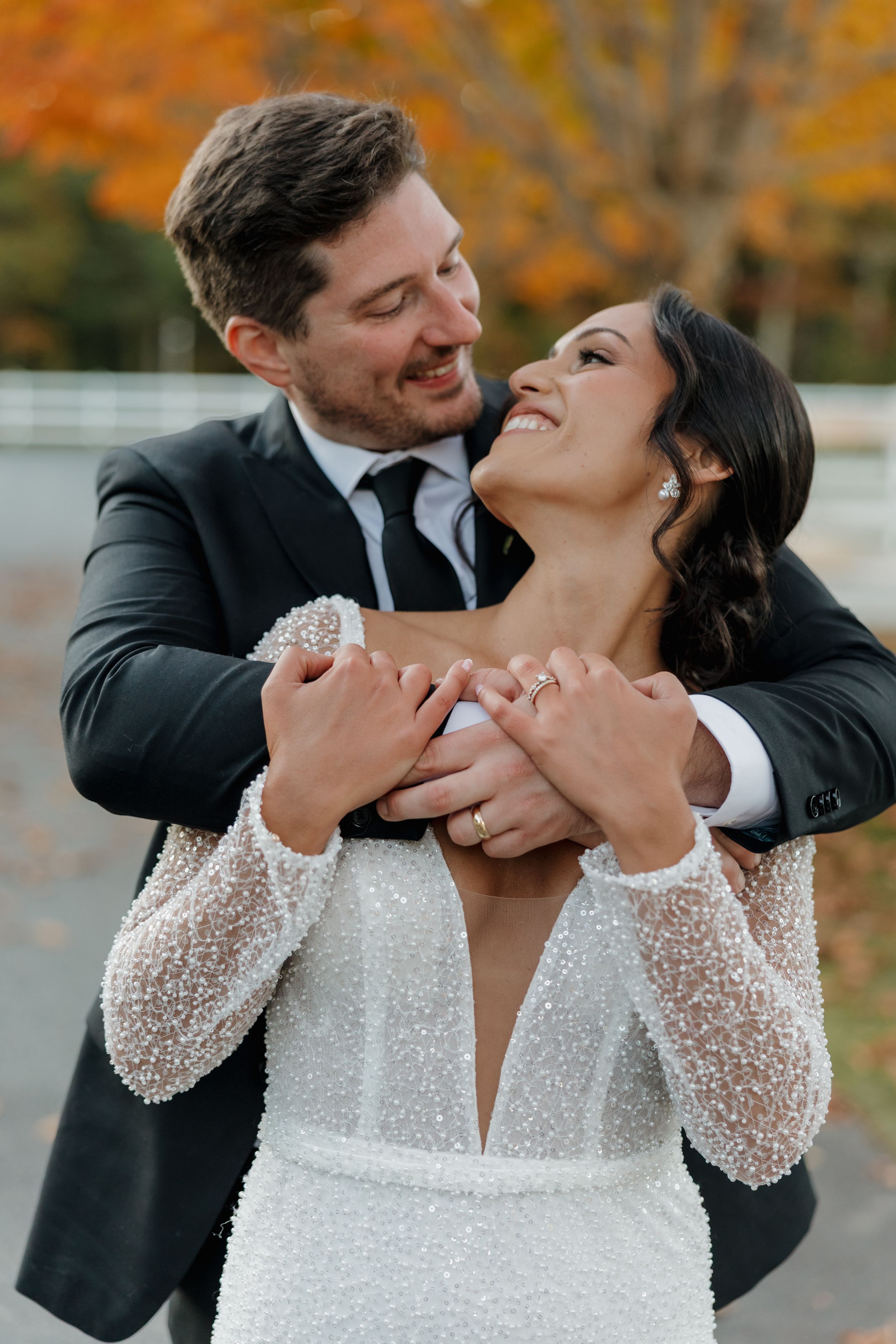 Bride and groom embrace joyfully outdoors in fall foliage, she in a pearl beaded gown, he in a dark suit with black tie.