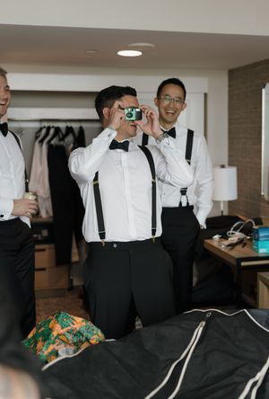 Groomsmen in white shirts and suspenders laugh as one photographs with a green camera in a hotel room.
