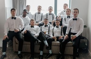 Group of groomsmen in white shirts, black bow ties, and suspenders posing together on a couch for a wedding photo.