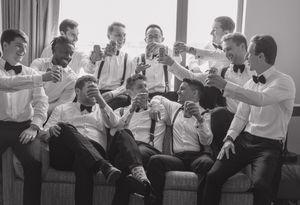 Black and white photo of groomsmen in bow ties toasting with drinks before a wedding ceremony.