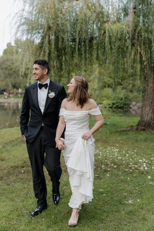 A bride and groom walk hand-in-hand by a pond, she in an off-shoulder gown, he in a black tuxedo.