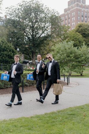 Three groomsmen in tuxedos carrying Bud Light cases walk through a park path on a wedding day.