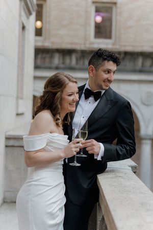 A bride in an off-shoulder white gown and groom in black tuxedo share a champagne toast on an elegant stone balcony.