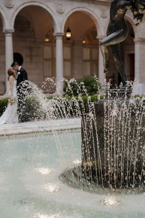 A couple shares a romantic kiss near an ornate fountain with a bronze statue at an elegant courtyard venue.