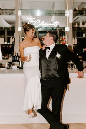 A couple shares a playful moment at a wedding reception bar with neon signage in the background.