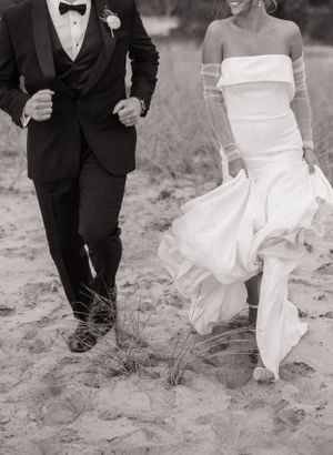 A couple in formal wedding attire walks together on sandy beach terrain in black and white.