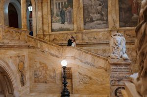 Couple sharing a romantic moment on grand marble staircase with ornate murals in an elegant historic building.