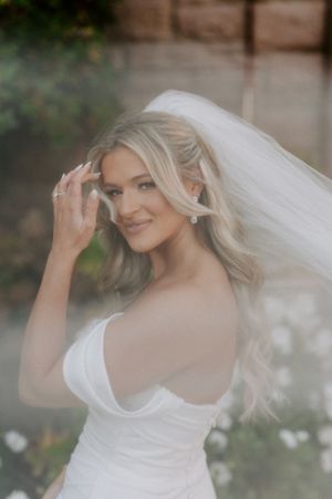 A bride in a white wedding dress adjusts her flowing veil against a blurred natural background.