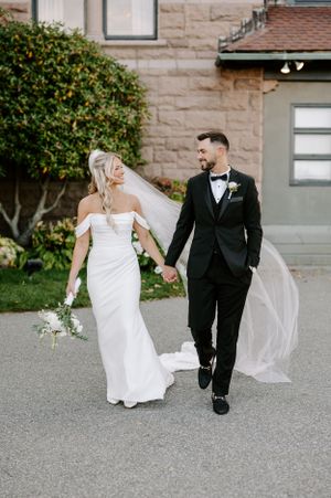 A couple in wedding attire walks together on a stone pathway beside a building.