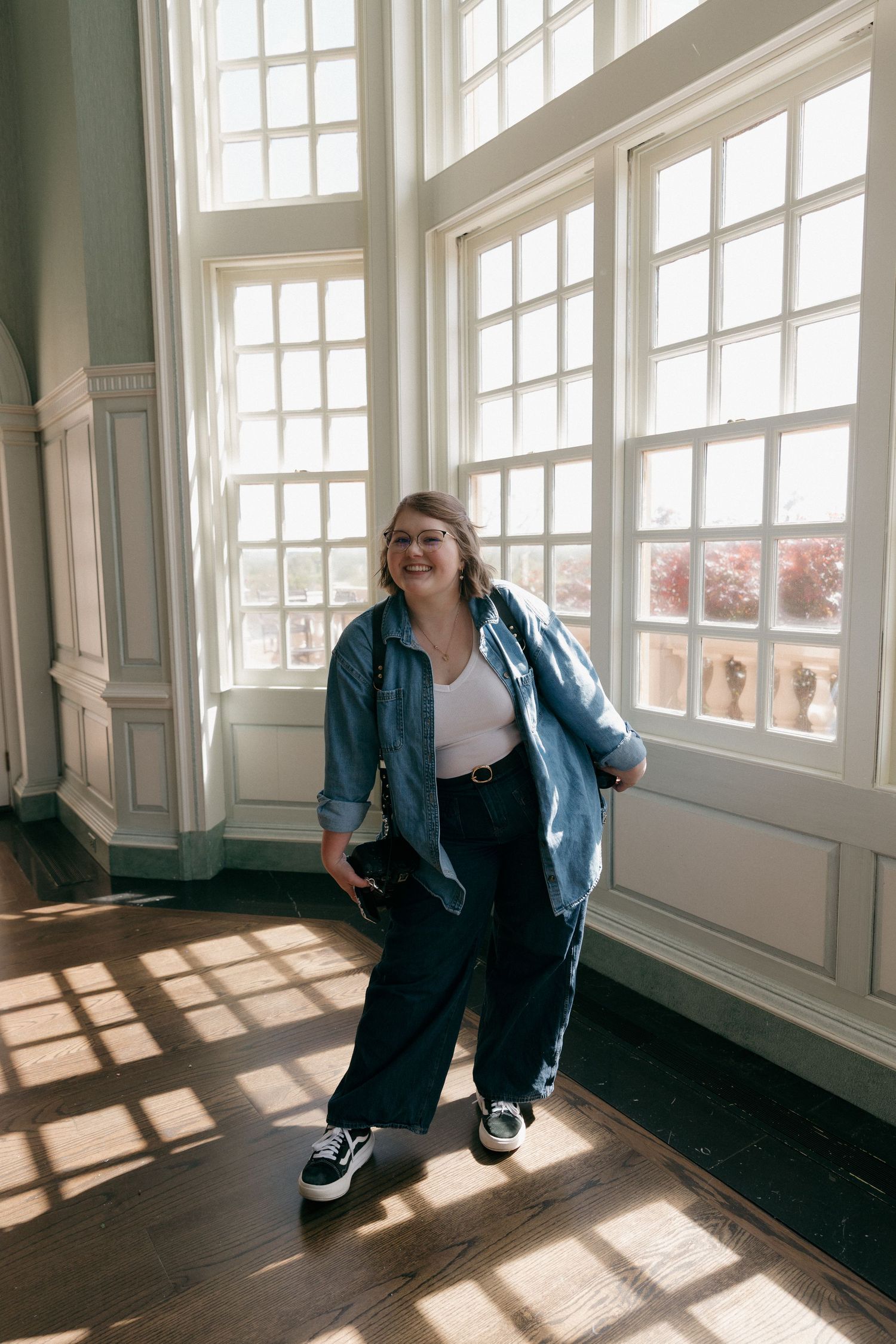 A person in a casual denim outfit stands near sunlit windows in an elegant room with hardwood floors and white trim.