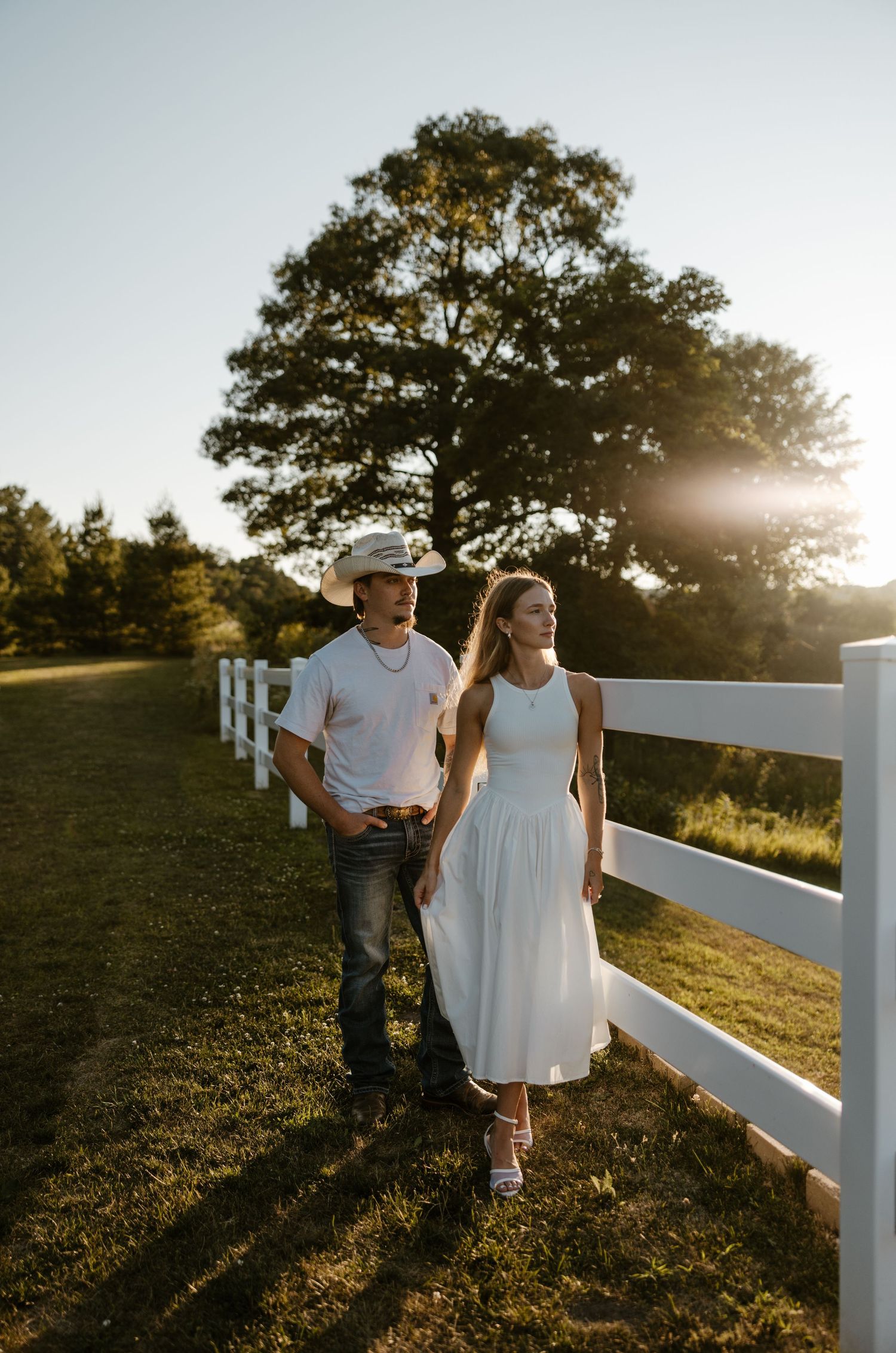 A young couple walks along a wooden fence on a grassy field with a tree in the background during a sunset. Fort Worth Texas photographer