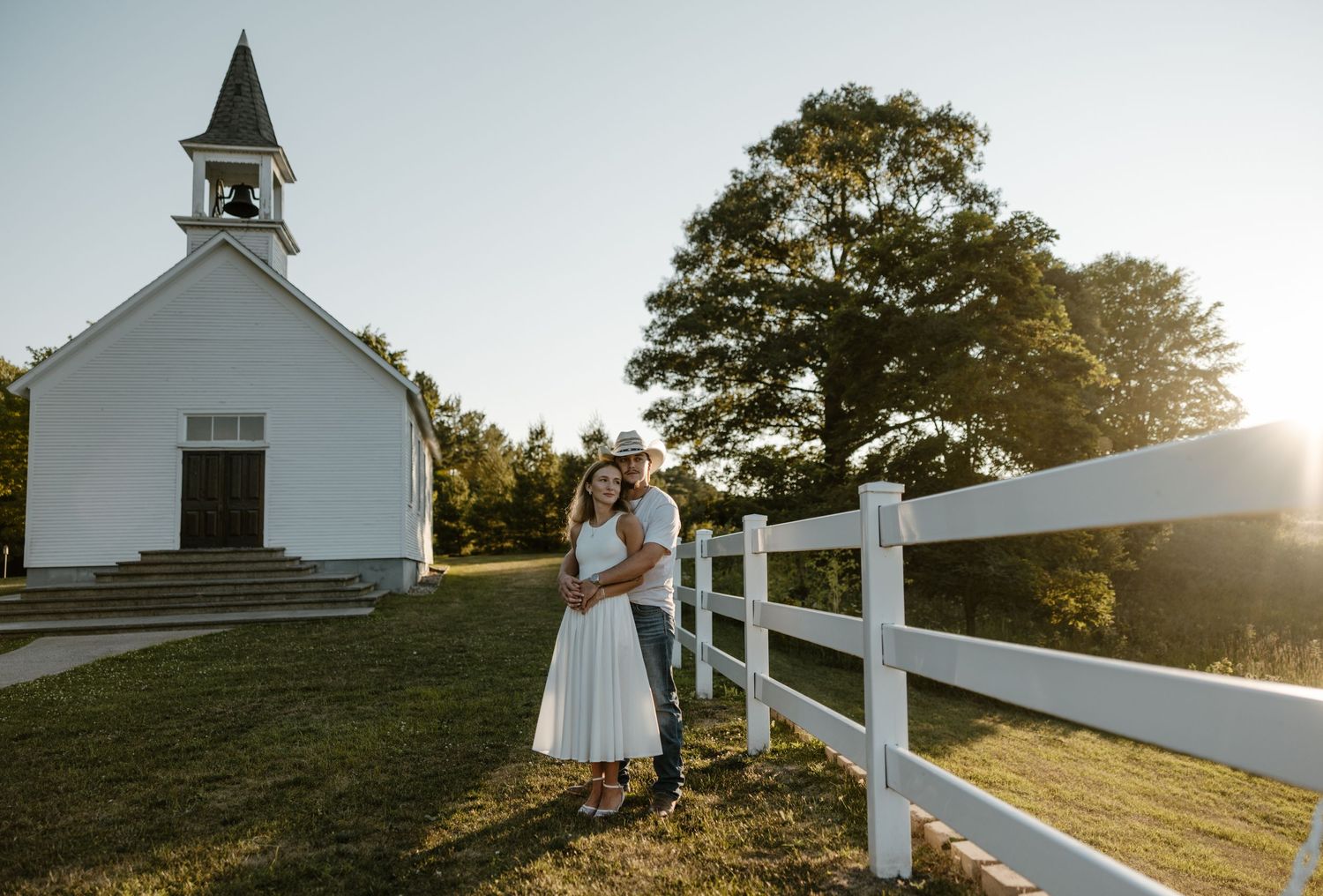 A bride and groom stand in front of a white church on a grassy field with a fence and trees in the background. Fort Worth Texas photographer