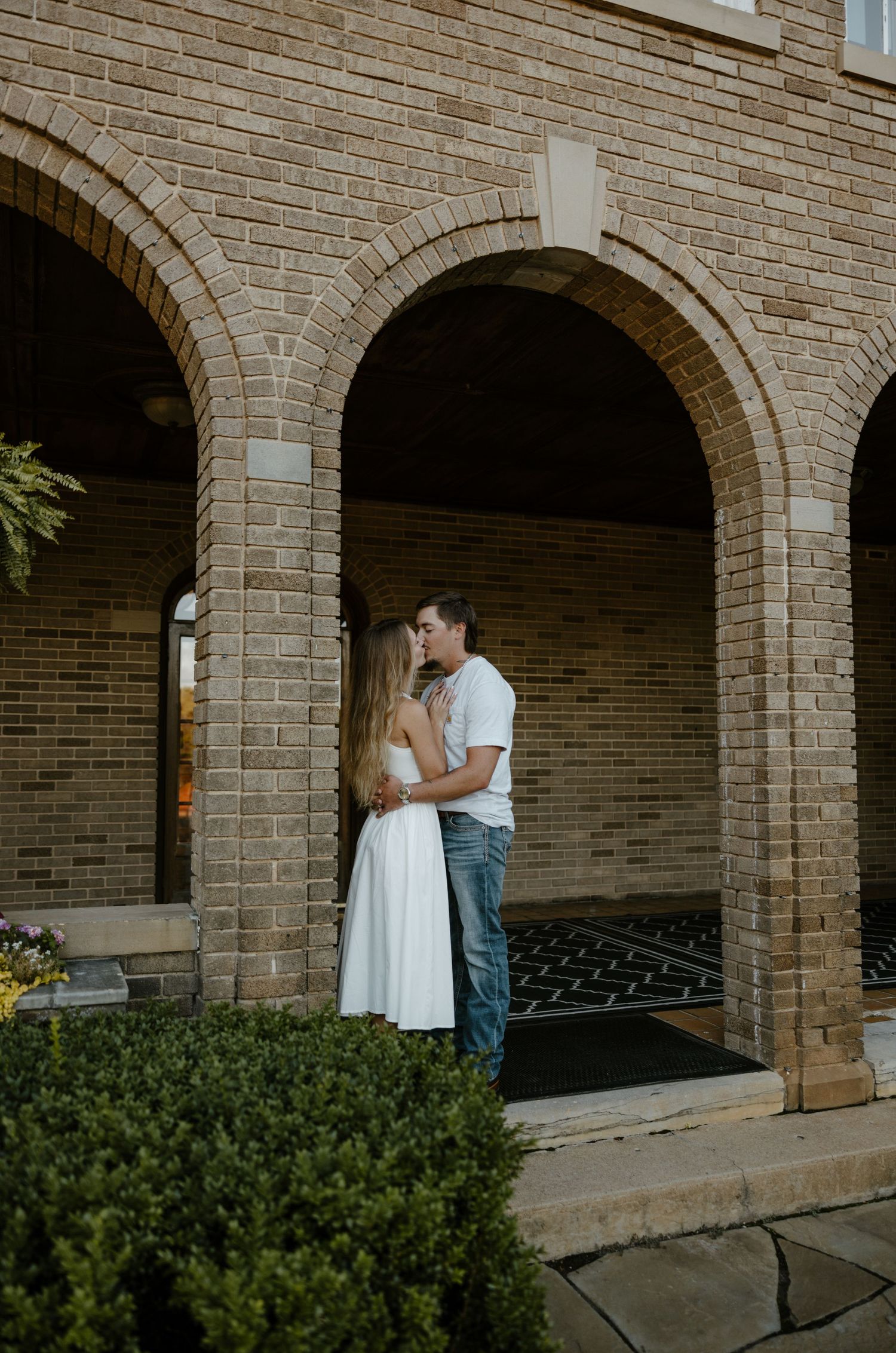 A couple embracing in front of a brick archway with greenery surrounding the entrance. Fort Worth Texas wedding photographer. Country photo