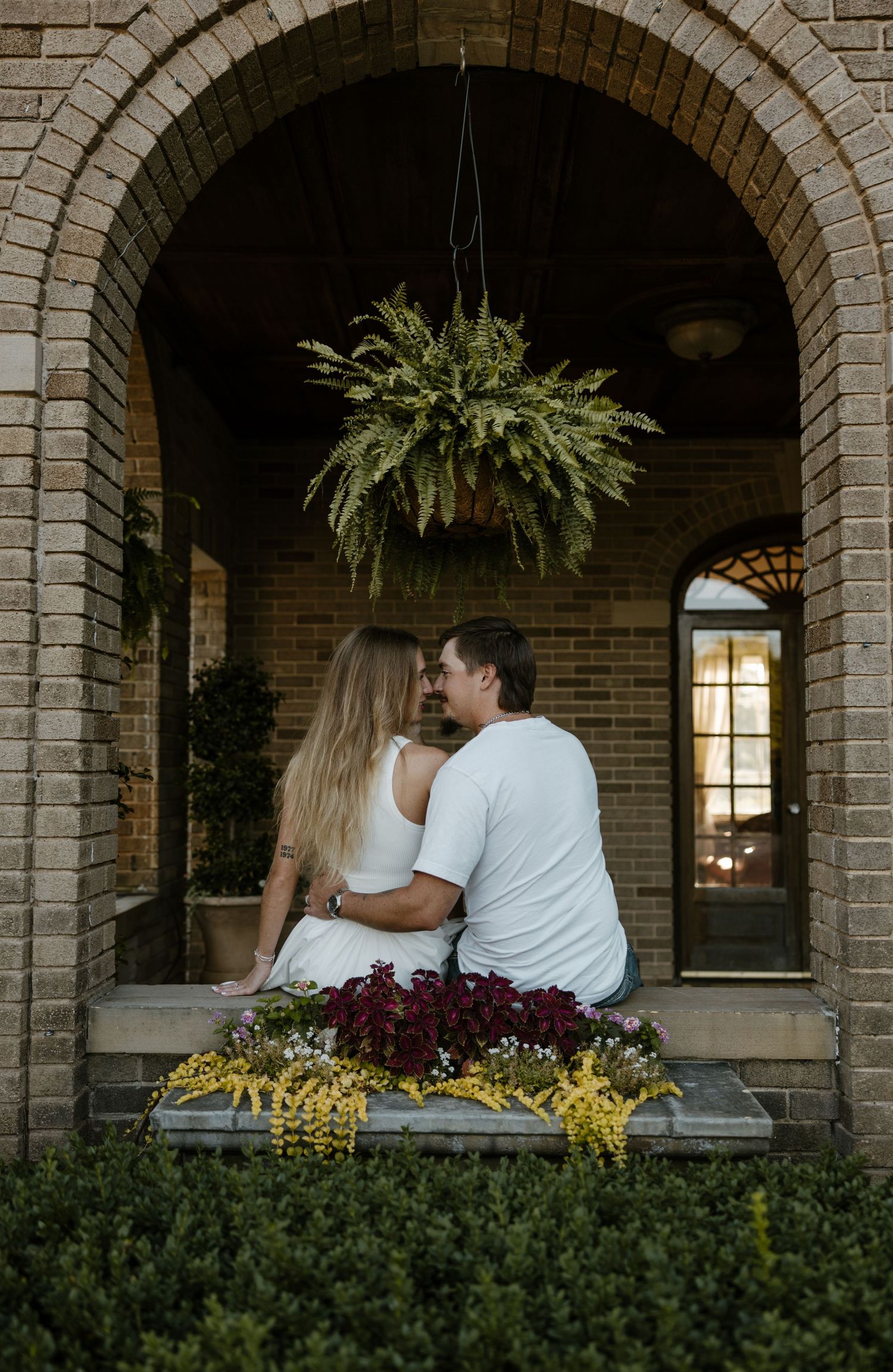 A young couple embracing in front of a brick archway, surrounded by lush greenery. Fort Worth Texas wedding photographer. Country photoshoot