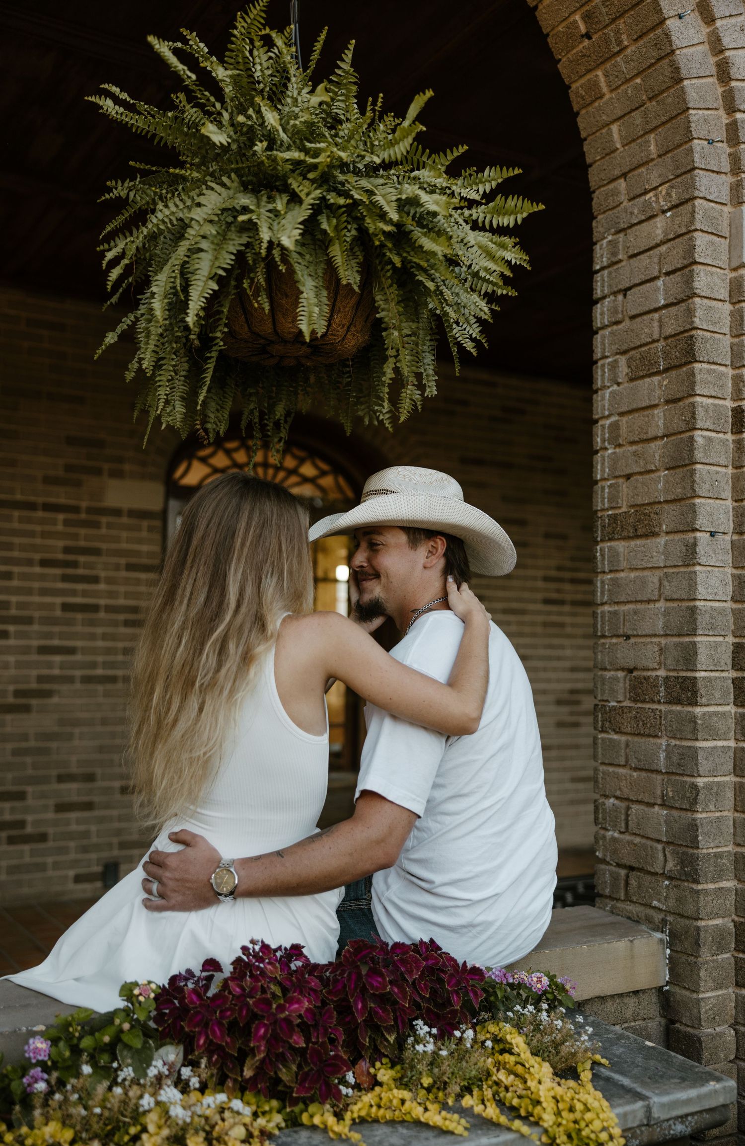 A couple embracing in front of a brick building with a large potted plant hanging above them. Fort Worth Texas wedding photographer.