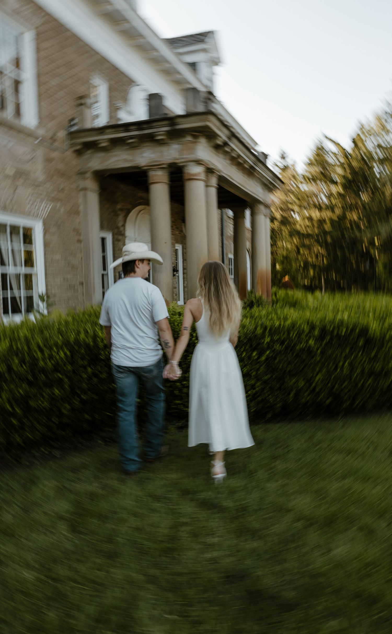 A couple walking hand-in-hand on a grassy path in front of a historic building with columns. Fort Worth Texas wedding photographer.