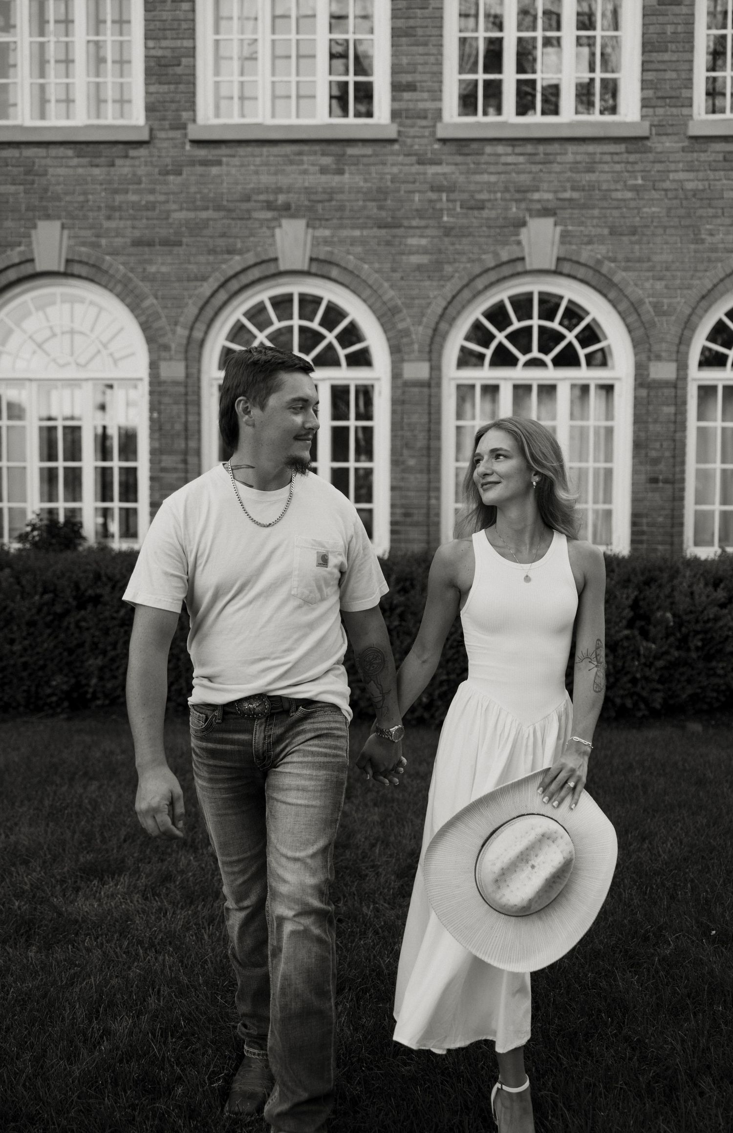 A black and white photograph shows a young couple walking together on a grassy lawn in front of a historic building with arched windows.