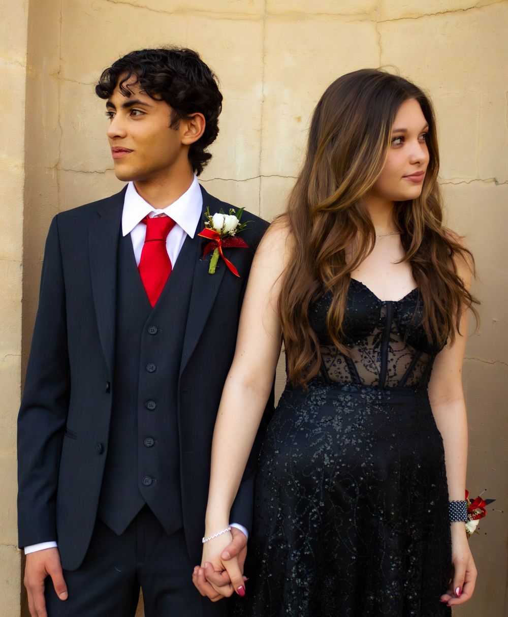 Prom couple in formal attire holding hands against a beige wall.