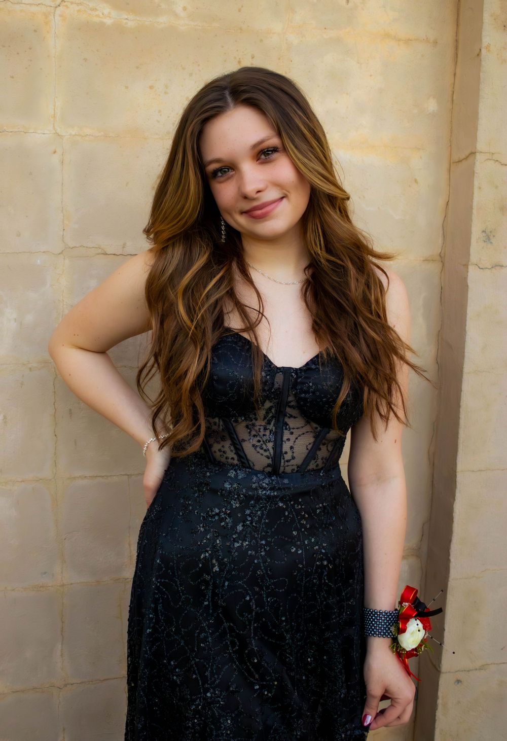 Young woman in black lace formal dress poses against beige wall.