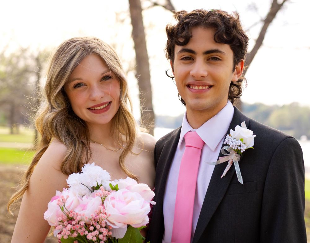 Couple in formal attire at prom, with pink flowers and boutonniere against an outdoor spring background.