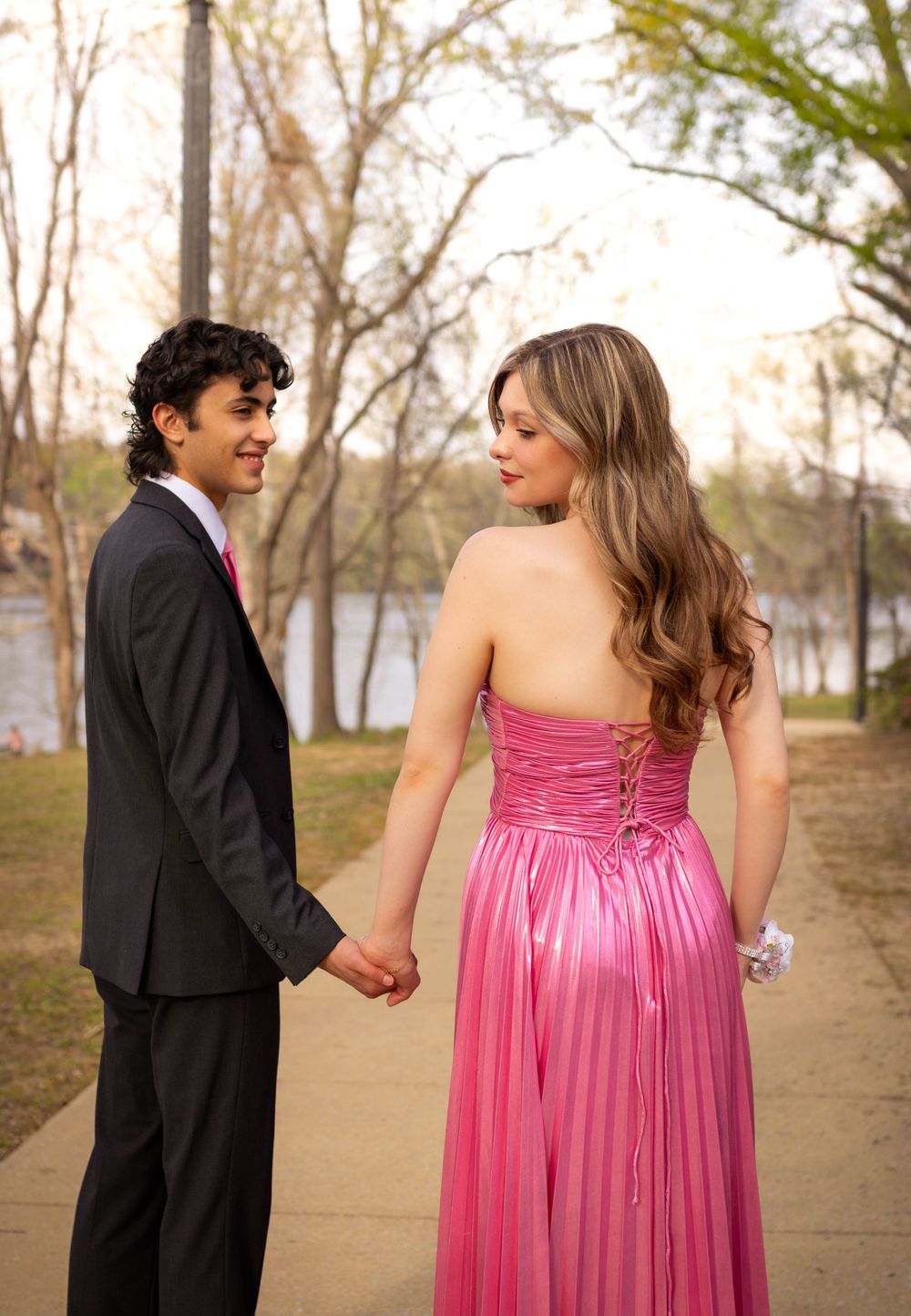 Couple in formal wear hold hands while walking along a path near a lake at sunset.