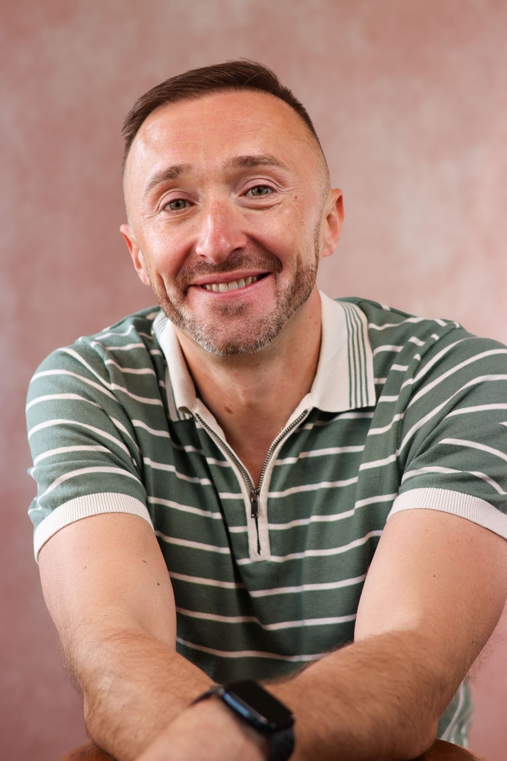 Middle-aged person in striped polo shirt smiling warmly against pink background.