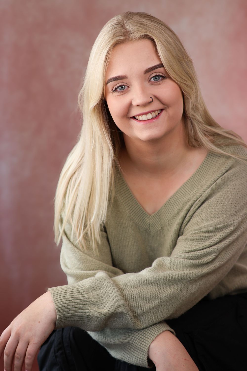 Person in olive v-neck sweater with long blonde hair sitting against pink background.
