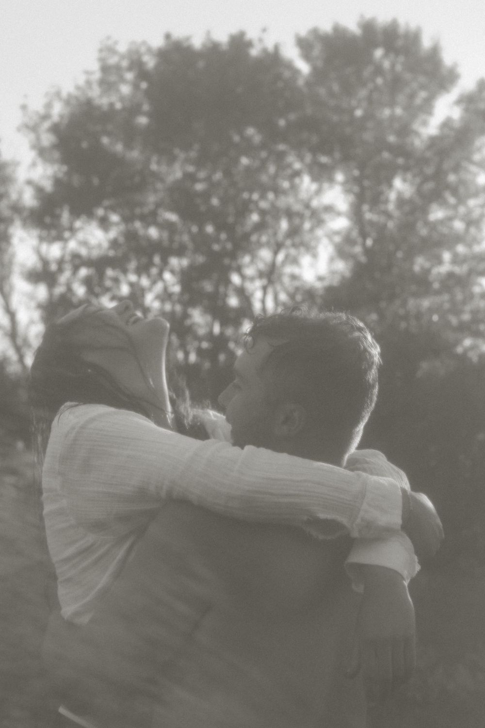 Romantic black and white portrait of a couple embracing outdoors with trees silhouetted in the background.