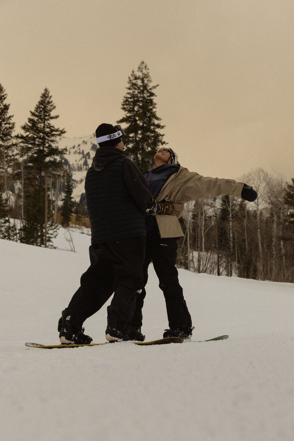 Snowboarders practicing techniques on a snowy slope with evergreen trees in background.