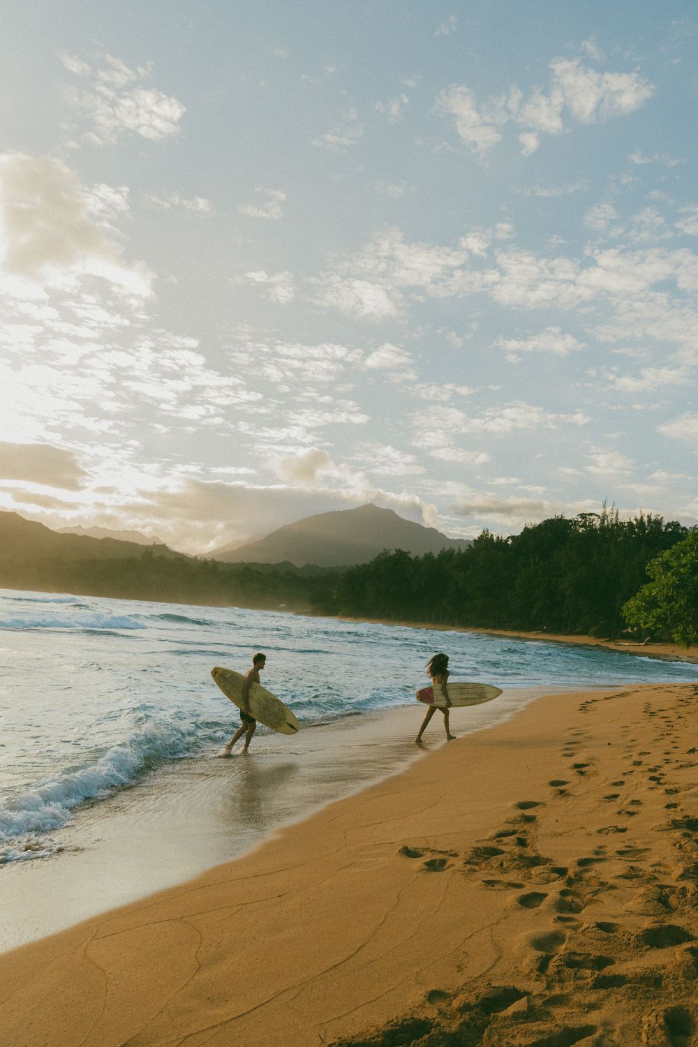 Surfers walking along a scenic beach at sunset with mountains in the background.