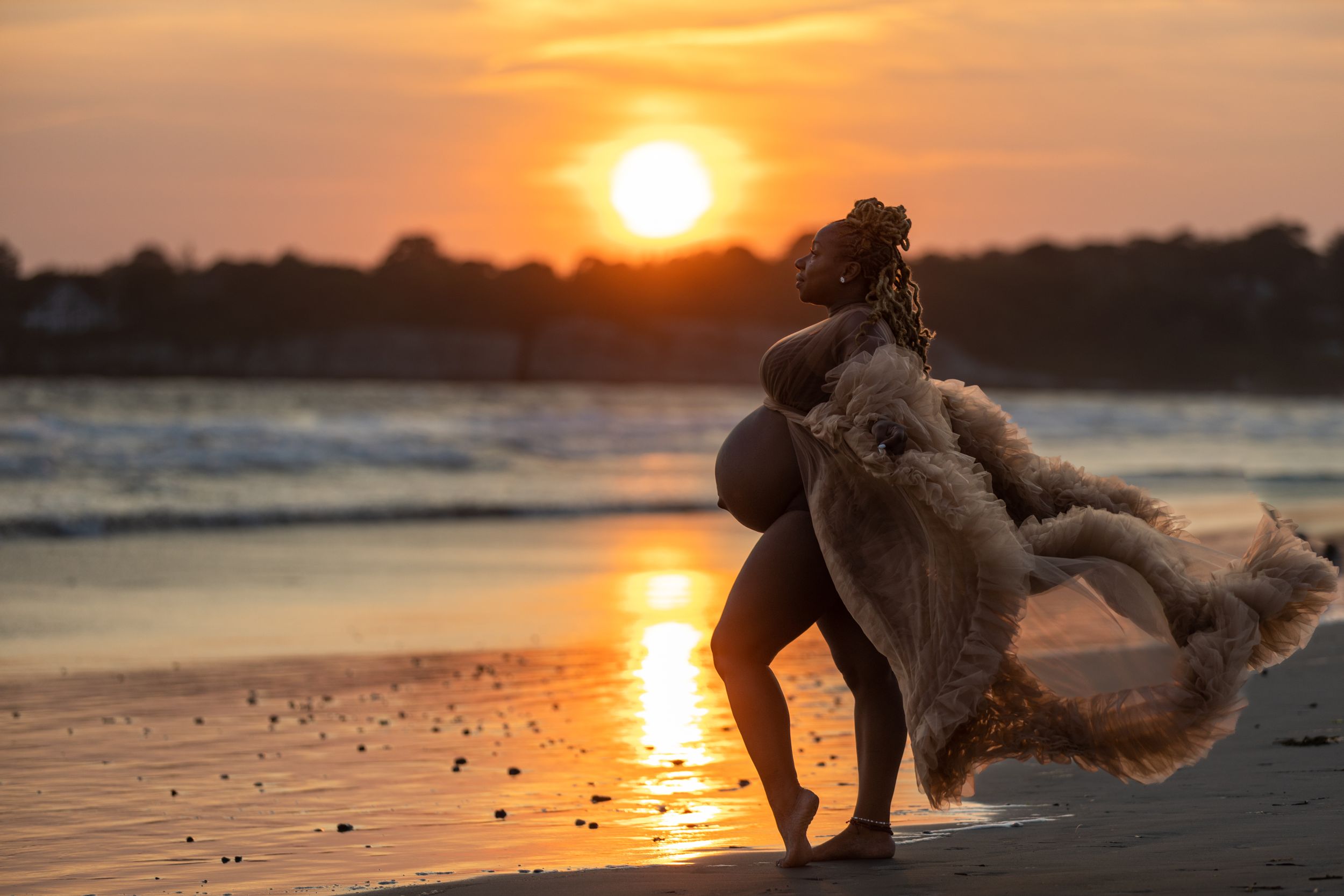 Silhouette of pregnant woman in flowing dress dancing on beach at golden sunset.